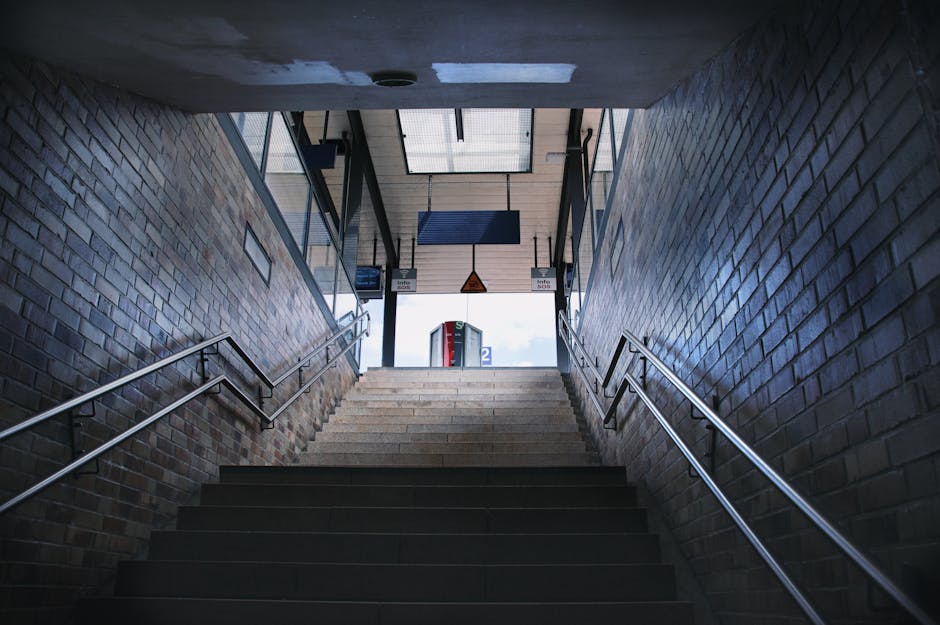 Photograph taken from the bottom of a staircase inside a building, showing a view upwards toward an exit with natural daylight. The staircase has dark steps with metal handrails on both sides, leading
